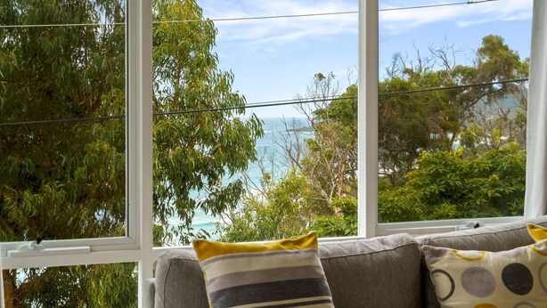 A view through large windows of a room with a grey sofa and cushions, overlooking a coastal area with trees and the sea. - Attunga - Relaxed family beach home among the gum trees (Wye River)