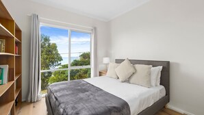 A bedroom with a large window, a bed with pillows, a bedside table with a lamp, and a bookshelf. - Attunga - Relaxed family beach home among the gum trees (Wye River)
