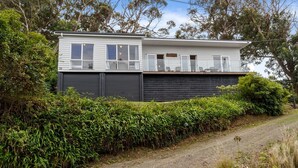 A two-story house with a dark exterior and white upper section, surrounded by dense greenery. - Attunga - Relaxed family beach home among the gum trees (Wye River)