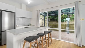 A modern kitchen with white cabinetry, a stainless steel refrigerator, a large island with bar stools, and a sliding glass door leading to an outdoor area. - Attunga - Relaxed family beach home among the gum trees (Wye River)