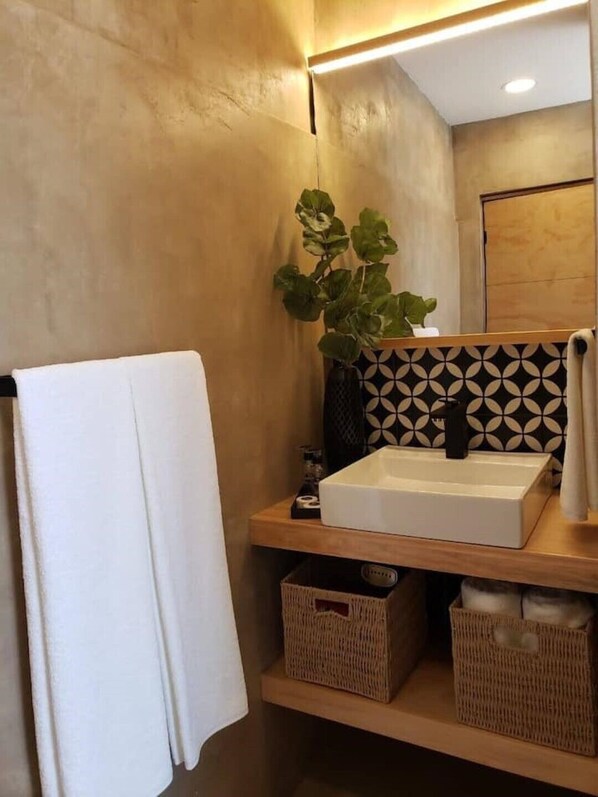 A bathroom with a white sink, a mirror, and a potted plant. There are two wicker baskets on the shelf below the sink.
