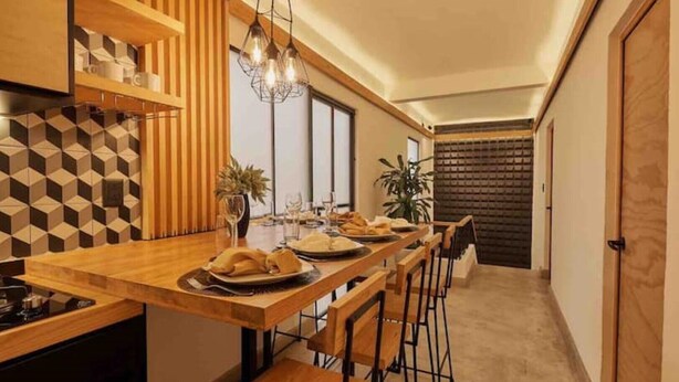 A modern kitchen with a wooden island, bar stools, and a dining table set for a meal. - Pinoli House, Xochimilco Neighborhood (Oaxaca)