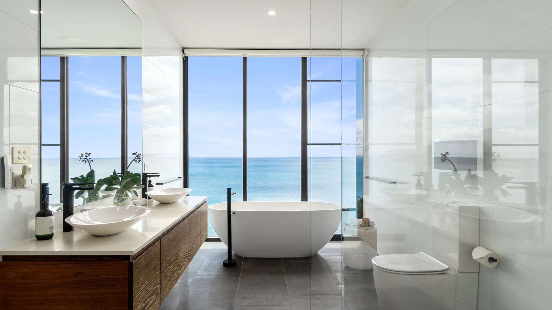 A modern bathroom with a freestanding bathtub, a large mirror, and a view of the sea.