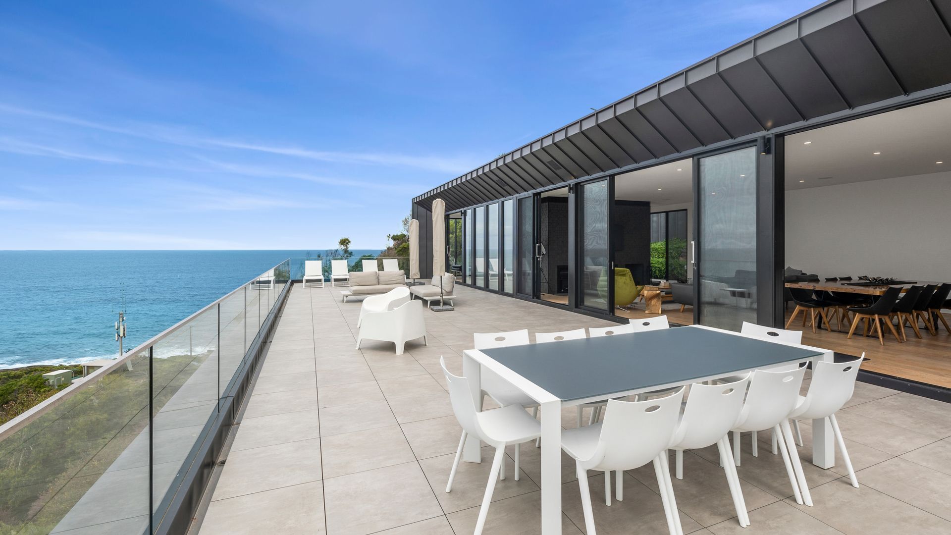 A modern outdoor patio with a view of the sea, featuring a dining area with white chairs and a table, and a covered seating area with umbrellas.