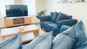 Ein Wohnzimmer mit einem Flachbildfernseher auf einem Holzboden, einem blauen Sofa mit vielen Kissen und einem Couchtisch.