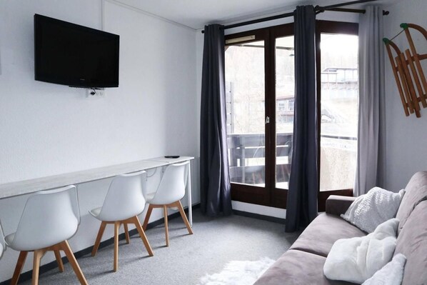 A modern living room with a sofa, a TV mounted on the wall, a dining area with chairs, and a sliding glass door leading to an outdoor area.