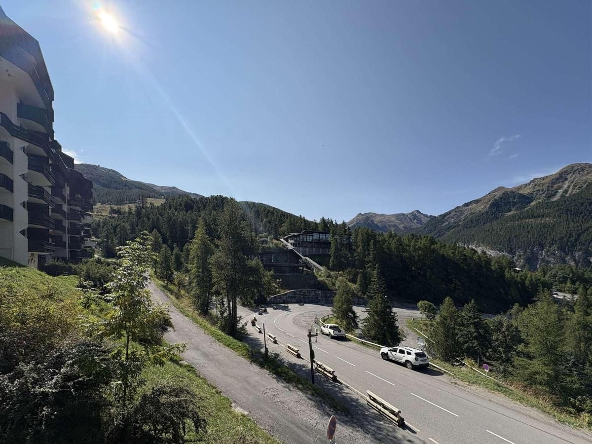 Une route sinueuse traverse une région montagneuse, avec un bâtiment sur la gauche et une colline boisée en arrière-plan.