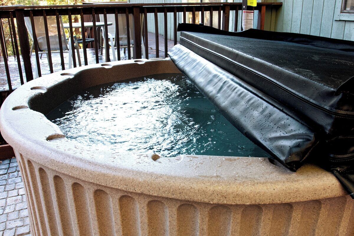 A hot tub with a stone base and a black cover, situated on a patio with a railing and outdoor furniture in the background.