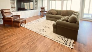 Un salón amplio con piso de madera, un sofá modular grande, una silla de madera y un televisor de pantalla plana montado en la pared.