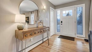 A hallway with a wooden console table, a round mirror, and a lamp. There is a door with a window and a doorbell. - Pickleball Palace! 3 Courts | Game Room | Hot Tub (Colorado Springs)