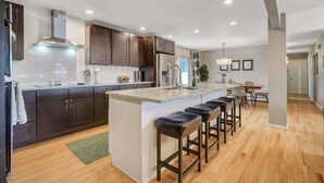 A modern kitchen with dark cabinets, a central island with bar stools, and a dining area in the background. - Pickleball Palace! 3 Courts | Game Room | Hot Tub (Colorado Springs)