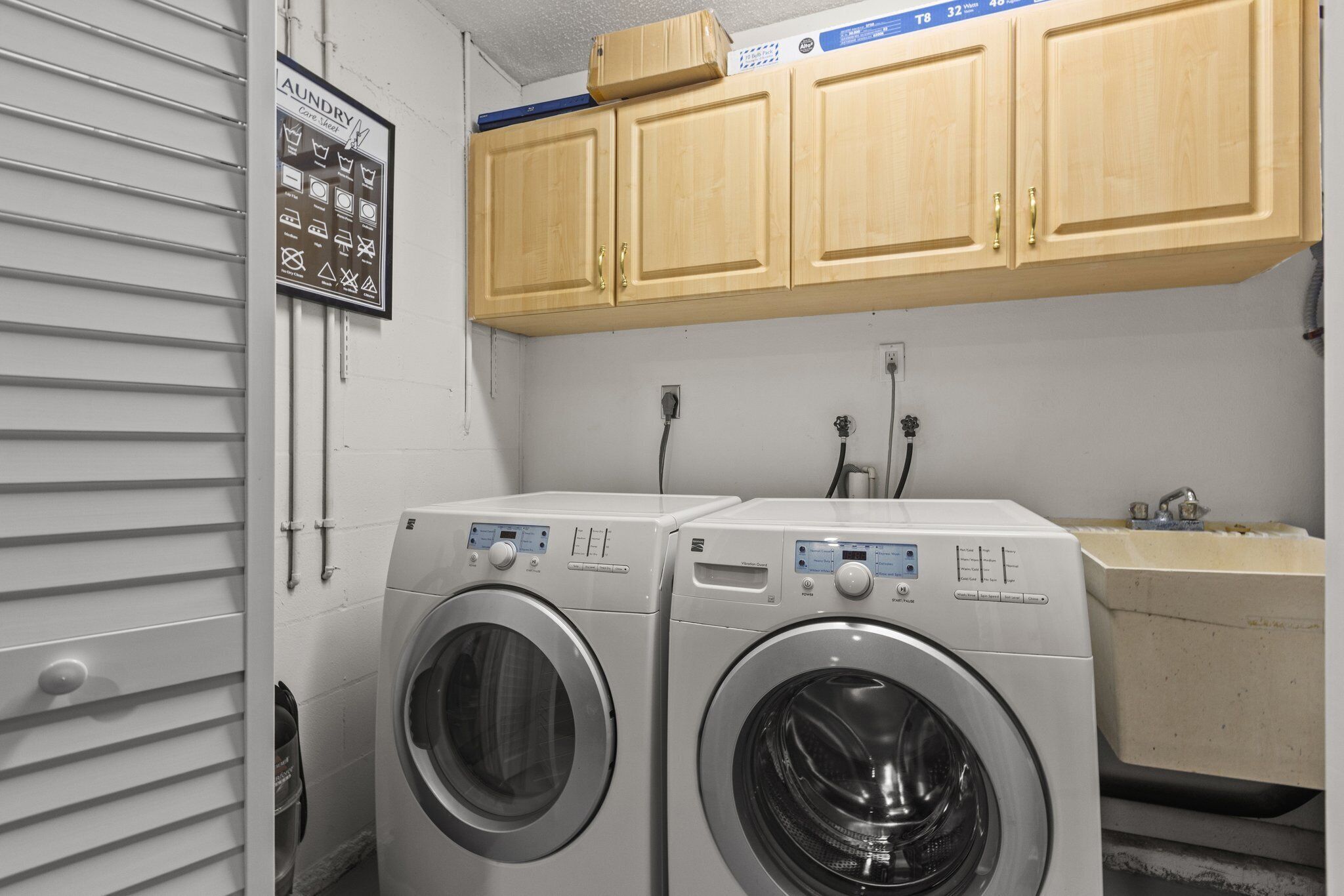 A laundry room with two front-loading washing machines, wooden cabinets above, and a sink with a faucet.
