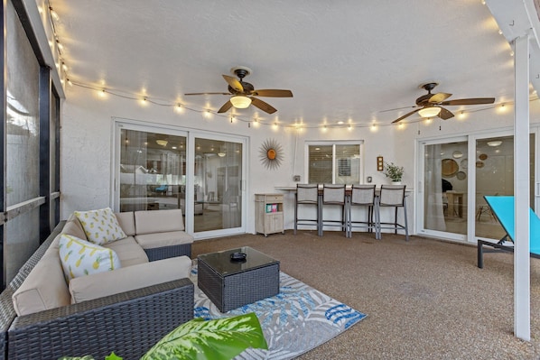 A covered patio area with a sofa, dining table, ceiling fans, string lights, and a sun decoration.