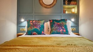 A bed with two colorful pillows, a round mirror above, and a shelf with books and a lamp. - The Jungle Nook: Romantic Hideaway with Log Burner (Berwick-upon-Tweed)