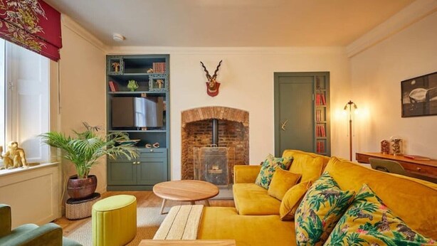 A cozy living room with a yellow sofa, a fireplace, a bookshelf, and a decorative antler on the wall. - The Jungle Nook: Romantic Hideaway with Log Burner (Berwick-upon-Tweed)