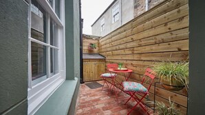 A small patio area with a brick floor, a wooden wall, and a window. There is a small table with chairs and potted plants. - The Jungle Nook: Romantic Hideaway with Log Burner (Berwick-upon-Tweed)