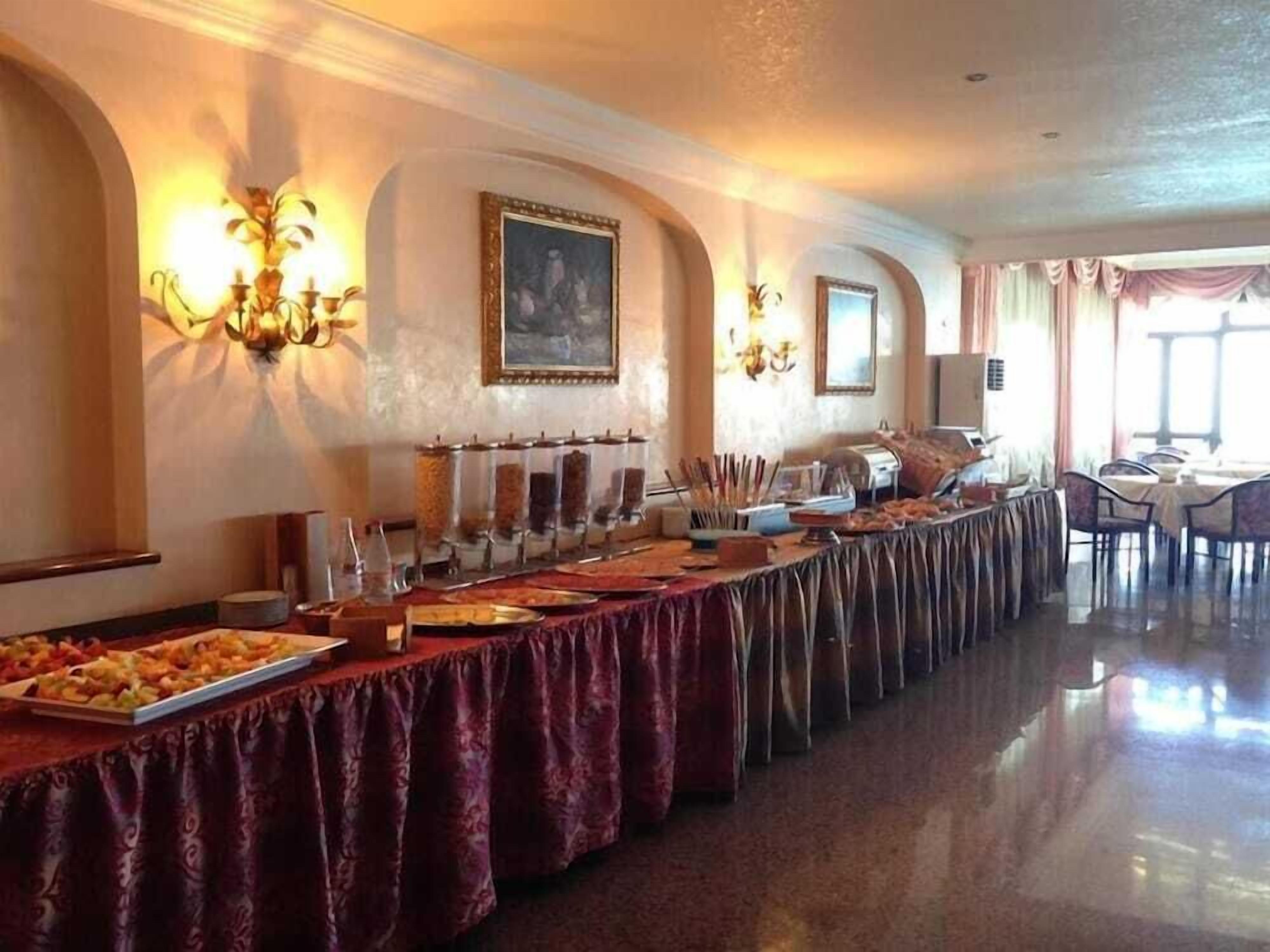Ein Festsaal mit einem langen Tisch, der mit einer roten Tischdecke bedeckt ist, Essensspendern und einem Gemälde an der Wand.