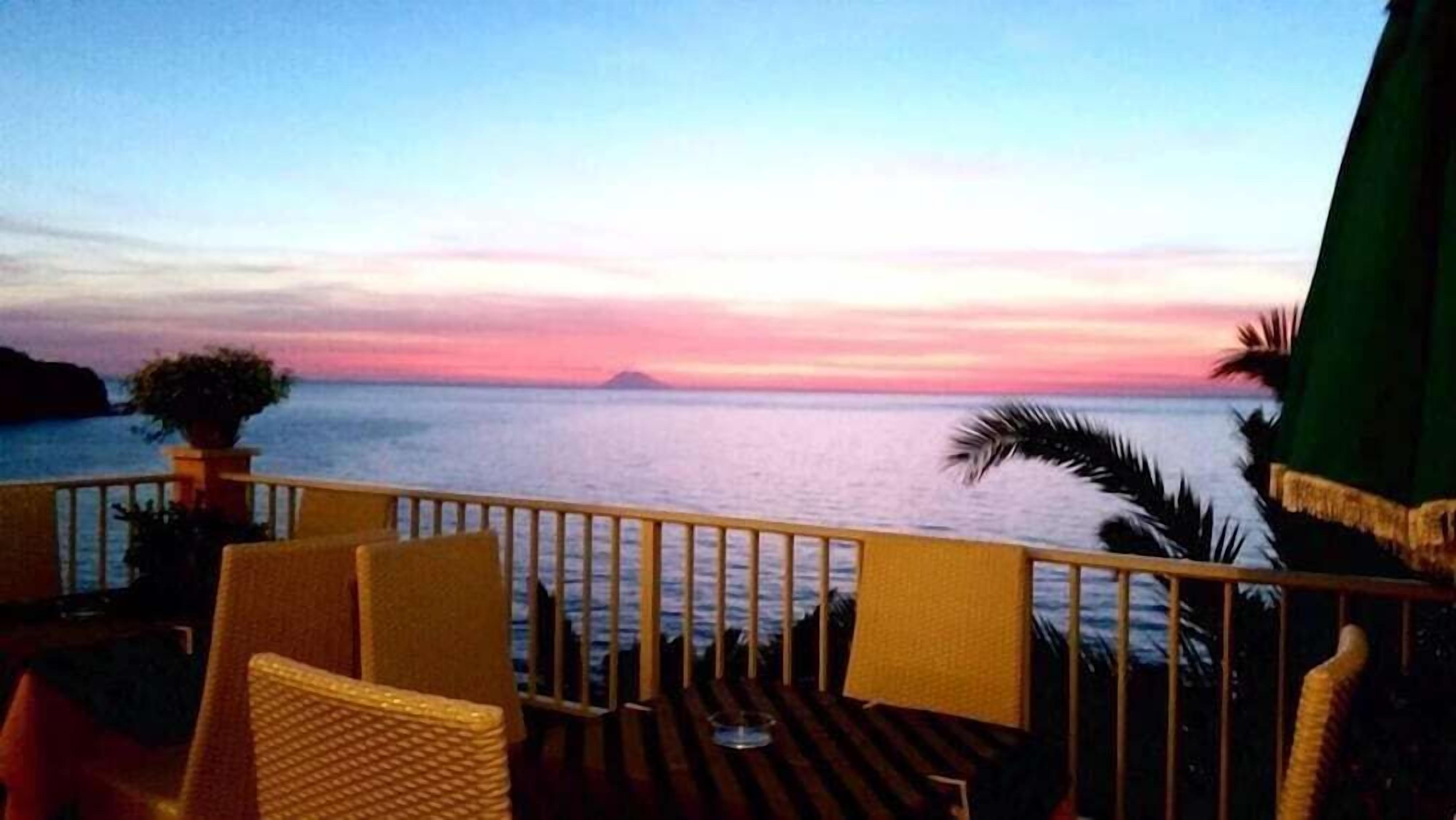 Eine Terrasse mit Korbstühlen und einem Tisch, mit Blick auf ein ruhiges Meer bei Sonnenuntergang.