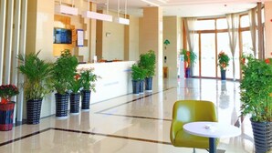 A hotel lobby with a reception desk, potted plants, a green chair, and a large window. - Greentree Inn Hefei Lujiang County No.1 Jin Street Express Hotel (Hefei)