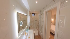 Een moderne badkamer met een marmeren wastafel, een grote spiegel, een douchecabine en een aangrenzende slaapkamer, zichtbaar door een open deur.