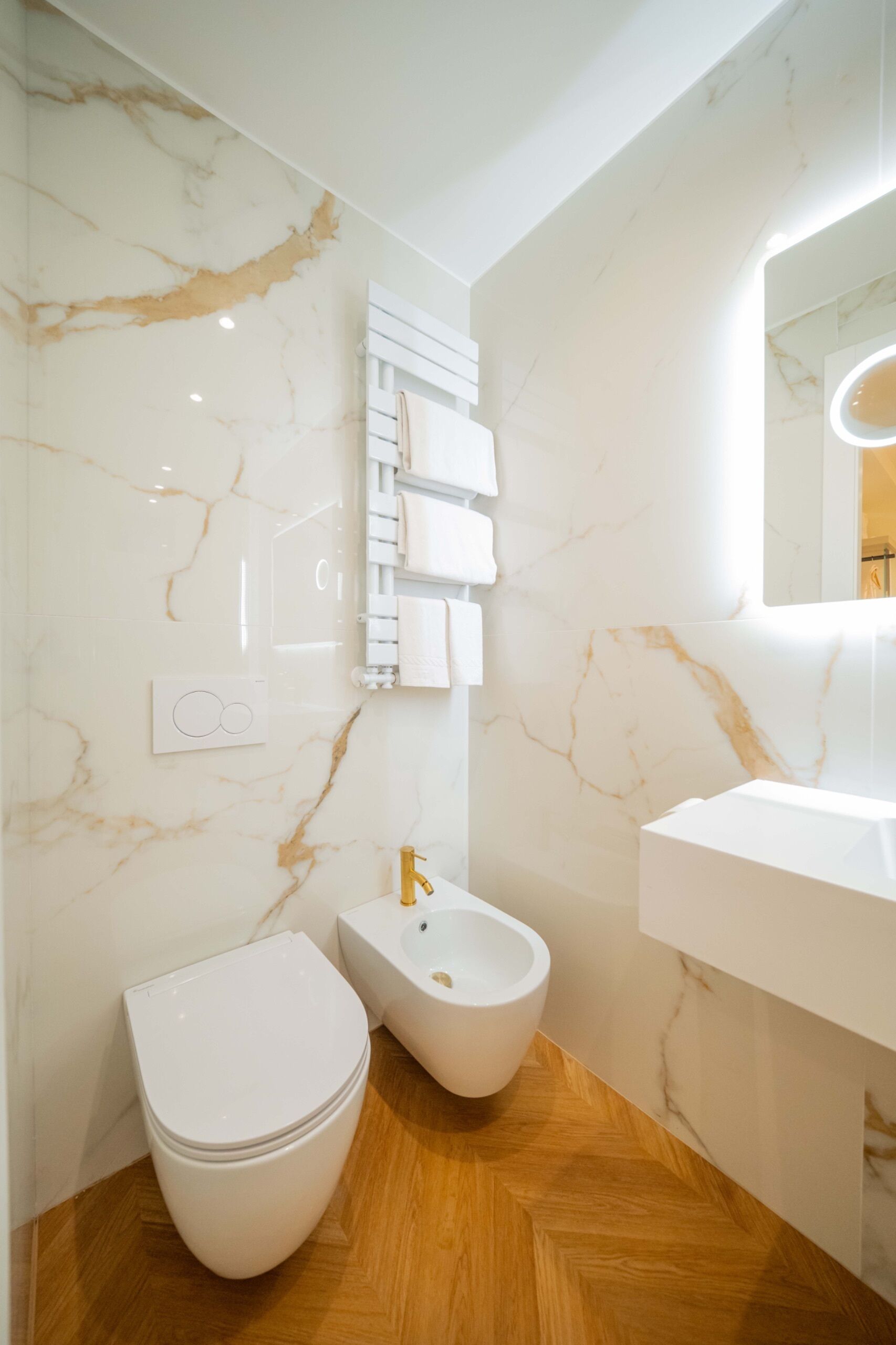 Un bagno con parete in marmo, pavimento in legno, WC bianco, bidet e lavabo con specchio.