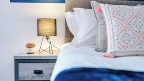 Ein ordentlich gemachtes Bett mit weißer Bettwäsche, einem dekorativen Kissen, einer Nachttischlampe und einem kleinen Nachttisch mit einem Glas.