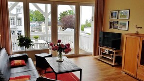 A living room with a sofa, a coffee table, a TV, and a cabinet. There is a balcony with a view of houses and trees. - Close to the beach and with sea views, apartment in a quiet, central location (Göhren)