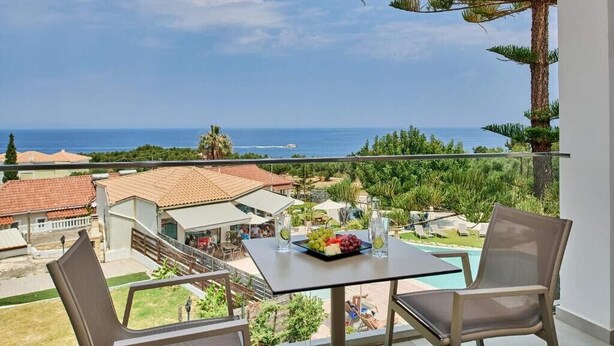 Un balcó amb taula i cadires, amb vista a una piscina i al mar i edificis.