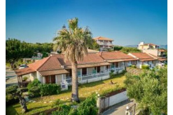 A residential area with houses featuring red-tiled roofs and white walls, surrounded by greenery and a clear blue sky. - Ionian Aura (Akrotiri)
