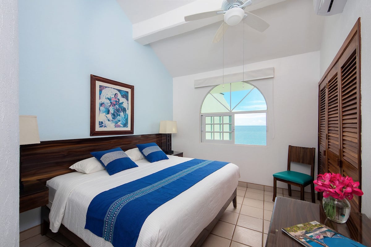 A neatly made bed with a blue and white bedspread, a wooden headboard, a nightstand with a lamp, a chair with a green cushion, a vase with pink flowers, and a window with a view of the sea.