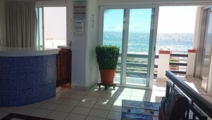 Zona de recepción de un hotel con mostrador de azulejos azules, una planta en maceta y vistas al mar a través de amplios ventanales.