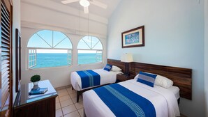 Habitación de hotel con dos camas, ventilador de techo, mesita de noche, lámpara, cuadro enmarcado y vistas al mar a través de amplias ventanas.