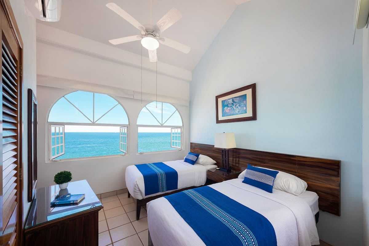 A hotel room with two beds, a ceiling fan, a nightstand, a lamp, a framed picture, and a view of the sea through large windows.