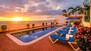 Un área junto a la piscina con sillones de descanso, una piscina y vistas al atardecer sobre el océano.