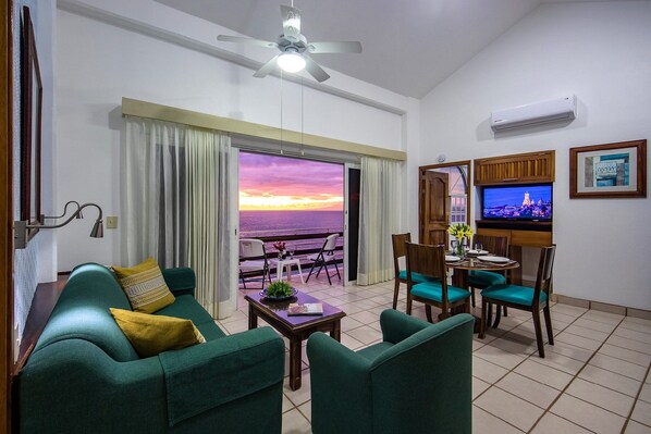 A living room with a sofa, chairs, a table, and a television. There is a view of the sunset through the window.