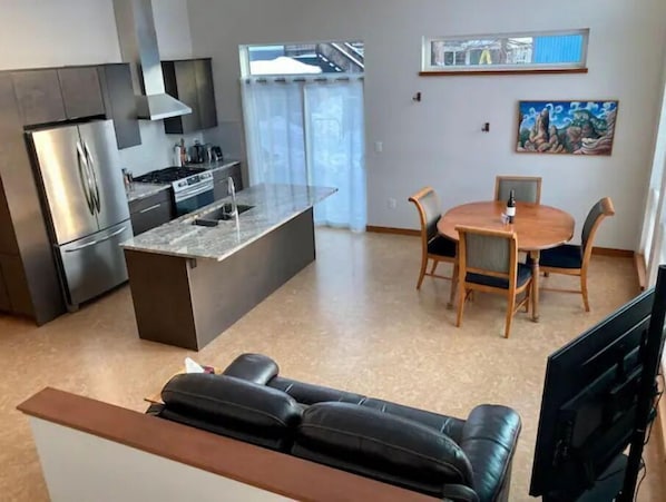 Una cuina moderna amb electrodomèstics d'acer inoxidable, una zona de menjador amb taula i cadires de fusta, i un sofà de pell negra al primer pla.