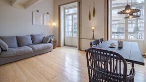 A living room with a sofa, a dining table with chairs, a lamp, and large windows with a view of buildings. - Porto Vitoria 5 in Oporto (Porto)
