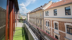A narrow balcony with a green artificial turf floor, bordered by a metal railing, overlooking adjacent buildings with traditional architecture. - Hotel Orion Varkert (Budapest)