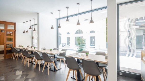 A modern dining area with long tables and chairs, large windows with a view of buildings, and pendant lighting. - Hotel Orion Varkert (Budapest)
