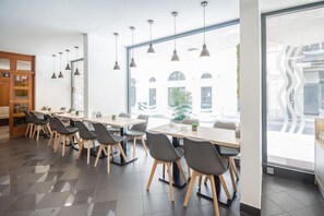 Ein moderner Essbereich mit langen Tischen und Stühlen, großen Fenstern mit Blick auf Gebäude und Pendelleuchten.
