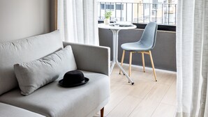A modern living room with a grey sofa, a black hat, a small table, and a chair. A balcony with a view of buildings is visible through the window.
