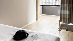 A hotel room with a large bed, a black hat on the bed, a balcony with a view of buildings, and a window with curtains.
