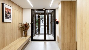 A modern hotel lobby with a wooden reception desk, a framed abstract artwork, a potted plant, and a glass door leading to the outside.