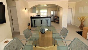 Un coin repas avec une table et des chaises en verre, une cuisine à l’arrière-plan et un téléviseur à écran plat fixé au mur.