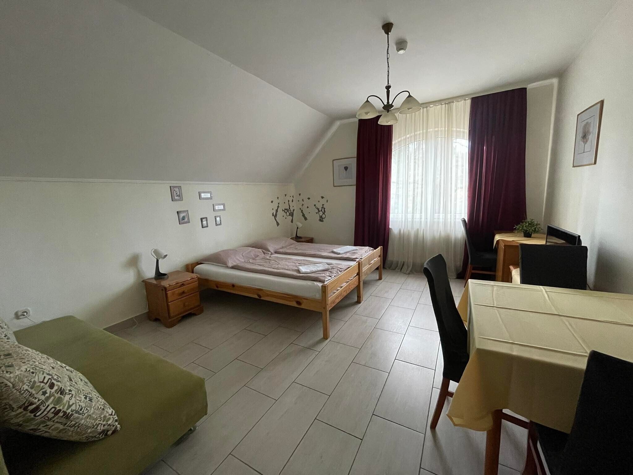 Una habitación pequeña y bien iluminada con cama, escritorio y silla. Hay cortinas en la ventana y un ventilador de techo.