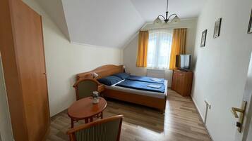 Un dormitorio con cama, una silla, una mesita, televisión y ventana con cortinas.