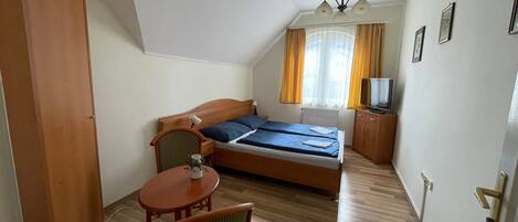 Un dormitorio con cama, una silla, una mesita, televisión y ventana con cortinas.