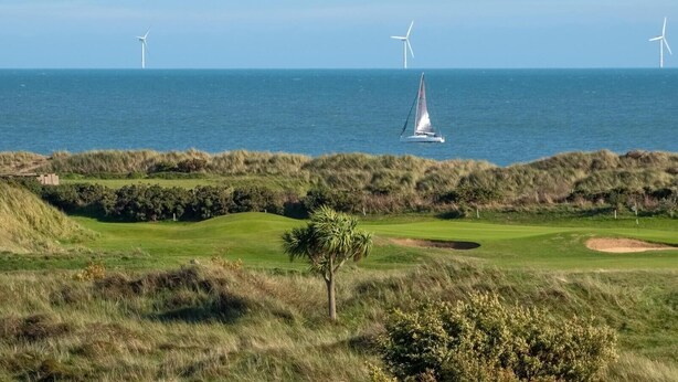 Un campo de golf costero con aerogeneradores al fondo y un velero en el agua.