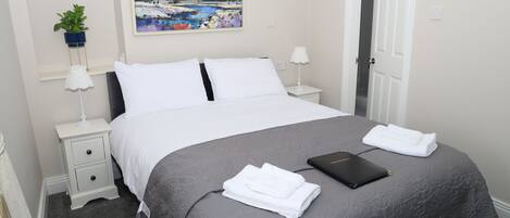 A neatly made bed with white linens, a gray quilt, and a black folder on the bed. There are two bedside tables with lamps and a potted plant on one.