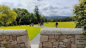 Puerta de piedra con la inscripción "The Wicklow Way" que da acceso a un parque con un sendero cubierto de césped y gente caminando.